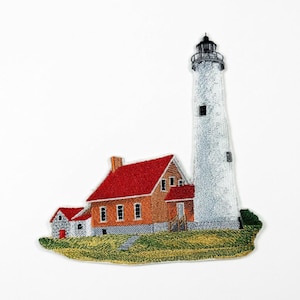 May include: An embroidered patch featuring a tall white lighthouse standing next to a house with a bright red roof and orange walls. The structure sits on a patch of green grass, with detailed stitching throughout the entire design.