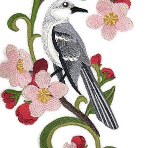 May include: An embroidered patch featuring a gray and white bird perched on a brown branch with green leaves, pink blossoms, and small red buds. The bird has a long, striped tail and a black beak, surrounded by floral elements in a vertical arrangement.