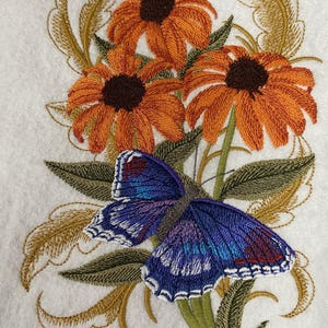 May include: Embroidered design featuring orange coneflowers with brown centers and a blue butterfly with white trim. The butterfly is positioned over green leaves and gold-colored foliage, all on a white background.