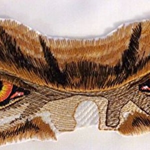 May include: Embroidered patch with a close-up of two yellow and orange eyes with black pupils. The eyes are set against a brown and white background, resembling a predator's face.