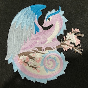 Peut inclure: Un patch brodé représentant un dragon rose et bleu avec des ailes, perché sur une branche avec des fleurs roses. La queue du dragon est enroulée en spirale.