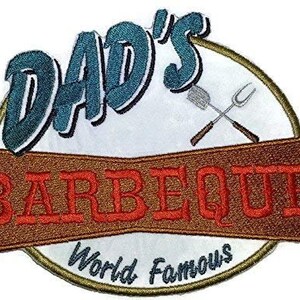 Może przedstawiać: Naszywka haftowana z brązowym tłem i tekstem "Dad's Barbeque World Famous" w czerwonych i białych literach. Naszywka zawiera również niebieski i biały napis "Dad's" w ozdobnym stylu pisma oraz parę skrzyżowanych narzędzi do grillowania.