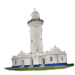 May include: An embroidered patch featuring a white lighthouse with a tall cylindrical tower and a base building. The structure has a blue foundation, dark gray railings, and a green grassy area at the bottom, all rendered in detailed thread work.