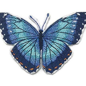 Puede incluir: Una mariposa azul con detalles negros y naranjas. La mariposa está bordada en una tela blanca.