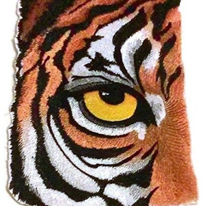 May include: A detailed patch featuring a tiger's eye and part of its face. The eye is yellow with a black pupil, surrounded by white, black, and orange striped fur. The patch has a textured appearance and a white border.