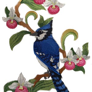 May include: Embroidered patch featuring a blue jay perched on a branch with white and pink orchids. The bird has detailed blue and white feathers. The branch has green leaves and gold accents. A decorative floral design.