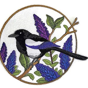 Peut inclure: Patch brodé représentant une pie noire et blanche perchée sur une branche avec des fleurs violettes et bleues. Le patch est circulaire et a un bord extérieur brun.
