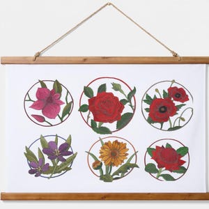May include: A white fabric wall hanging with a wooden frame and a jute rope hanger. The print features six circular illustrations of various flowers, including a pink clematis, a red rose, red poppies, purple crocuses, a yellow daisy, and a red protea.