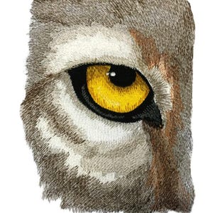 May include: An embroidered patch featuring a detailed animal eye with a bright yellow iris and a black pupil. The surrounding fur is rendered in shades of brown, tan, white, and gray thread, creating a textured appearance with visible stitching.