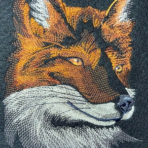 Könnte beinhalten: Gestickter Fuchs-Kopf mit orangefarbenem, braunem und weißem Fell. Der Fuchs schaut nach rechts mit einem leichten Lächeln. Der Hintergrund ist ein dunkelgrauer Stoff.