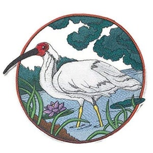 Puede incluir: Parche bordado con un ibis blanco con cabeza y pico rojos de pie en un estanque con una flor morada. El ibis está rodeado de juncos verdes y el fondo es un estanque azul con un árbol verde.
