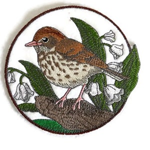 Peut inclure: Patch brodé représentant un oiseau brun et blanc perché sur une branche avec des feuilles vertes et des fleurs blanches. L'oiseau est entouré de fleurs de muguet.