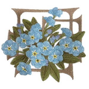 Op de afbeelding: Een geborduurde patch met een tros lichtblauwe vergeet-mij-nietjes met gele harten en groene bladeren, geplaatst over een beige, vierkant frame met een open midden en decoratieve, puntige hoekaccenten.