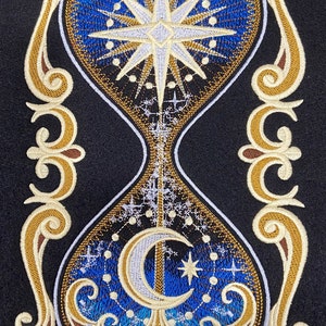 May include: An embroidered hourglass with a gold frame and a blue background. The top of the hourglass features a white star with a gold outline. The bottom of the hourglass features a white crescent moon with a gold outline. The hourglass is surrounded by a gold decorative frame.