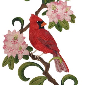 May include: Embroidered patch featuring a red cardinal perched on a branch with pink flowers. The cardinal is detailed with black eyes and a black beak. The flowers are pink with white centers. The branch is brown with green leaves.