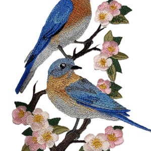 May include: Embroidered patch featuring two bluebirds perched on a branch with pink and yellow flowers. The birds have blue and brown feathers, with detailed stitching. The patch is on a white background.