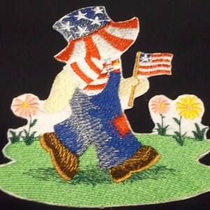 May include: Embroidered iron-on patch of a child wearing overalls and a red, white, and blue hat holding a small American flag. The child is standing in a green grassy field with two flowers.