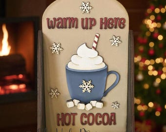 以下が含まれることがあります： 「warm up here」と「Hot Cocoa」の文字が入った木製看板。泡立てたクリームと雪の結晶が入った青いマグカップが特徴です。看板には「SVG Laser File」の文字も含まれています。