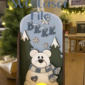 May include: A wooden winter decoration with a polar bear wearing a scarf, snowflakes, and trees. The design includes the text "SVG Laser File BRRR" and a warm light. The design is from Elea Veda Designs.