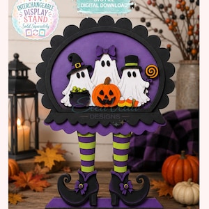 Könnte beinhalten: Ein mehrschichtiges Halloween-Display aus Holz mit drei weißen Geistern, einem Kürbis und einem Kessel in einem gewellten schwarzen Rahmen. Die Geister haben grün-lila gestreifte Beine und schwarze Stiefel. Ein grüner Sockel zeigt den Text Boo Crew.