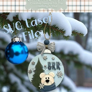Puede incluir: Adorno navideño redondo azul claro con diseño de oso, copos de nieve y la palabra "BRR". El adorno cuelga de una rama de pino nevada. También se ve una bola de cristal azul.