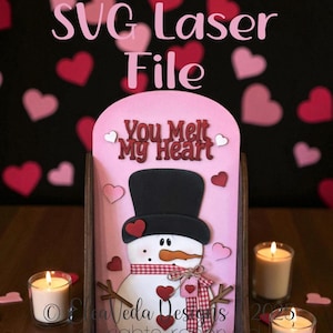 以下が含まれることがあります： 黒い帽子、ハートのアクセント、そして「You Melt My Heart」のテキストが入った雪だるまが特徴の、ピンク色の木製SVGレーザーファイル。デザインは、ハートの切り抜きと点灯したキャンドルを備えた黒い背景に設定されています。