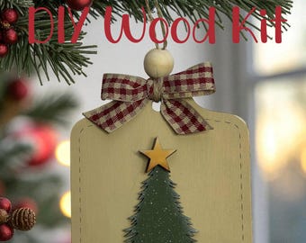 May include: A wooden Christmas tree ornament with a green tree and gold star design. The ornament has a beige background, a red and white checkered bow, and the text "DIY Wood Kit". The ornament is hanging from a Christmas tree branch.