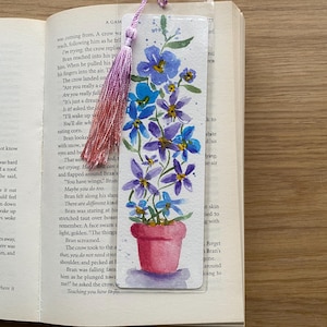 May include: A watercolour bookmark showing a pink flower pot with blue and purple flowers. A pink tassel hangs from the top. The bookmark is in an open book.
