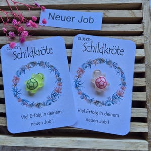 Könnte beinhalten: Zwei Karten mit Schildkröten-Anhängern, eine neongrün und eine rosa. Die Karten tragen das deutsche Wort "Schildkröte" und den Satz "Viel Erfolg in deinem neuen Job!" Auf der oberen Karte steht "Neuer Job."
