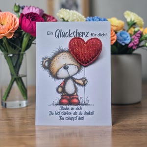 Könnte beinhalten: Eine Grußkarte mit einem Cartoon-Teddybären, der einen roten herzförmigen Ballon hält. Die Karte enthält deutschen Text, der sich mit "Ein Glücksherz für dich!" und inspirierenden Sätzen übersetzt. Bunte Blumen sind im Hintergrund.