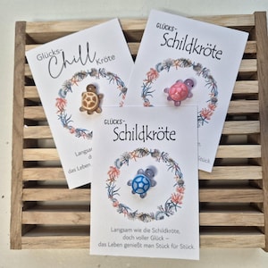 Könnte beinhalten: Drei Karten mit Schildkröten-Anhängern. Auf den Karten stehen die Wörter "Glücks-Schildkröte" und "Glücks-Chill Kröte". Die Anhänger sind braun, rosa und blau, jeweils umgeben von einem Kranz aus Muscheln und Korallen.