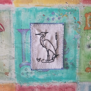 May include: A white fabric patch with a black embroidered image of a heron standing in water. The patch is surrounded by a blue denim border.