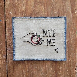 May include: A white fabric patch with blue denim edging, embroidered with a hand holding a red apple and the text "Bite Me" with a small heart.