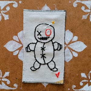 May include: Embroidered patch with a voodoo doll design. The doll is stitched in black thread on white fabric, with a red eye and a pin. The rectangular patch has a decorative border and is set against a brown background with white floral accents.