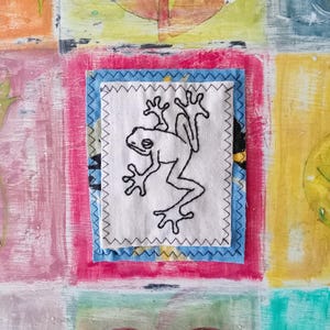 May include: A white fabric square with a black embroidered frog design. The square is framed with blue and yellow fabric borders.