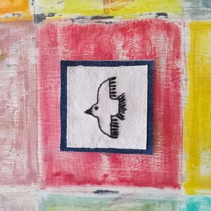 May include: A white fabric square with a black embroidered bird design, framed with a dark blue denim border. The square is displayed against a colorful painted background.