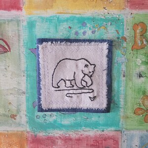May include: A white fabric square with a black embroidered image of a bear walking. The fabric is framed with a blue denim border.