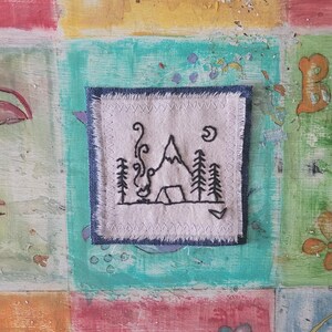 May include: A small embroidered patch featuring a black mountain, a tent, a campfire, and trees on a white background. The patch is edged with a blue denim border.