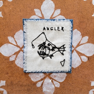 Peut inclure: Écusson carré en tissu avec un motif de poisson pêcheur noir cousu à la main et le mot « ANGLER » en lettres noires. Le patch a un fond blanc avec une bordure bleu denim effilochée.