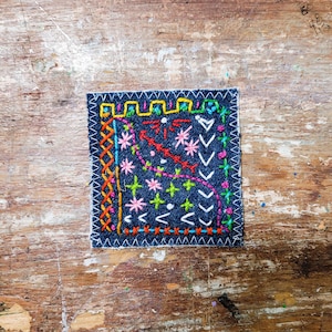 May include: A small square piece of denim fabric with colourful embroidery. The embroidery includes various stitches, such as cross-stitches, running stitches, and backstitches. The colours used are red, orange, yellow, green, blue, and pink.