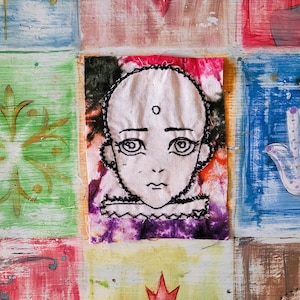 May include: A hand-stitched embroidered portrait of a face with large eyes and a small mouth. The portrait is on a colourful fabric background with purple, pink, orange, and green hues.