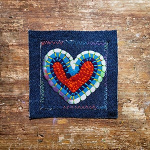 May include: A colourful, handcrafted textile heart design on a dark blue denim square. The heart is filled with red, blue, and yellow stitching, with decorative stitching around the edges. A charming piece of textile art.