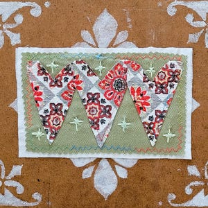 May include: A handmade textile art piece featuring three heart shapes with a floral pattern in red, black, and white. The hearts are set against a green background, stitched onto a white fabric, and framed by a brown background with white decorative elements.