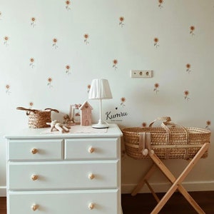May include: White chest of drawers with four drawers and wooden knobs. A wicker Moses basket with a white pom-pom sits on a wooden stand. The wall behind the dresser is decorated with small sunflower decals. The text "Kumru Home Design" is visible in the image.