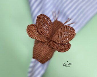 以下が含まれることがあります： 青と白のストライプのナプキンに置かれた、茶色の編まれた蝶のナプキンリング。ナプキンリングは自然素材で作られており、素朴な外観をしています。