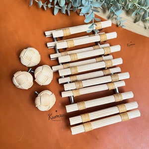 Può includere: Un set di 10 tiretti per cassetti in bambù intrecciato naturale con finitura marrone chiaro. I tiretti sono disposti in fila su una superficie in pelle marrone. Ci sono anche quattro pomelli in bambù intrecciato più piccoli sulla superficie.