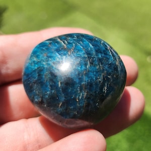 May include: A polished, rounded gemstone in shades of deep teal and blue, with subtle brown veining. The stone is held in a hand, showcasing its smooth, reflective surface. The gemstone's color and texture are highlighted against a blurred green background.