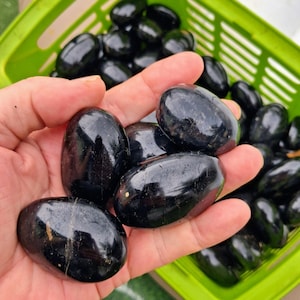 Op de afbeelding: Een hand die verschillende gepolijste, zwarte stenen vasthoudt. De stenen zijn glad, ovaal van vorm en hebben een glanzende afwerking. Een groene mand gevuld met vergelijkbare stenen is op de achtergrond. Deze stenen kunnen voor decoratieve doeleinden of als onderdeel van een verzameling worden gebruikt.