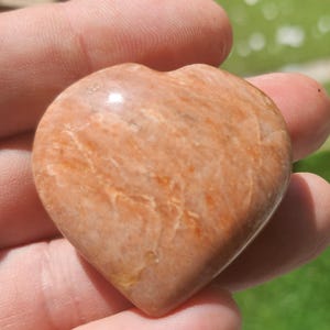 Può includere: Una pietra a forma di cuore con una superficie lucida. La pietra è di un caldo colore pesca con sottili variazioni di tono e venature. Il cuore è tenuto in una mano, con uno sfondo verde sfocato.