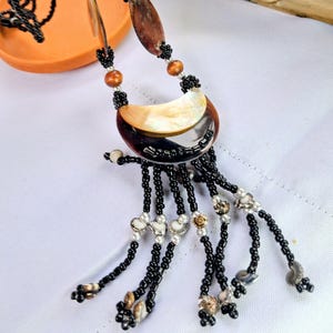 Peut inclure: Un collier artisanal avec un pendentif en forme de croissant et une incrustation de nacre. Le collier comporte des brins de perles noires avec des accents décoratifs, notamment de petites perles et des breloques en forme de coquillage. Le collier est présenté sur une surface blanche.